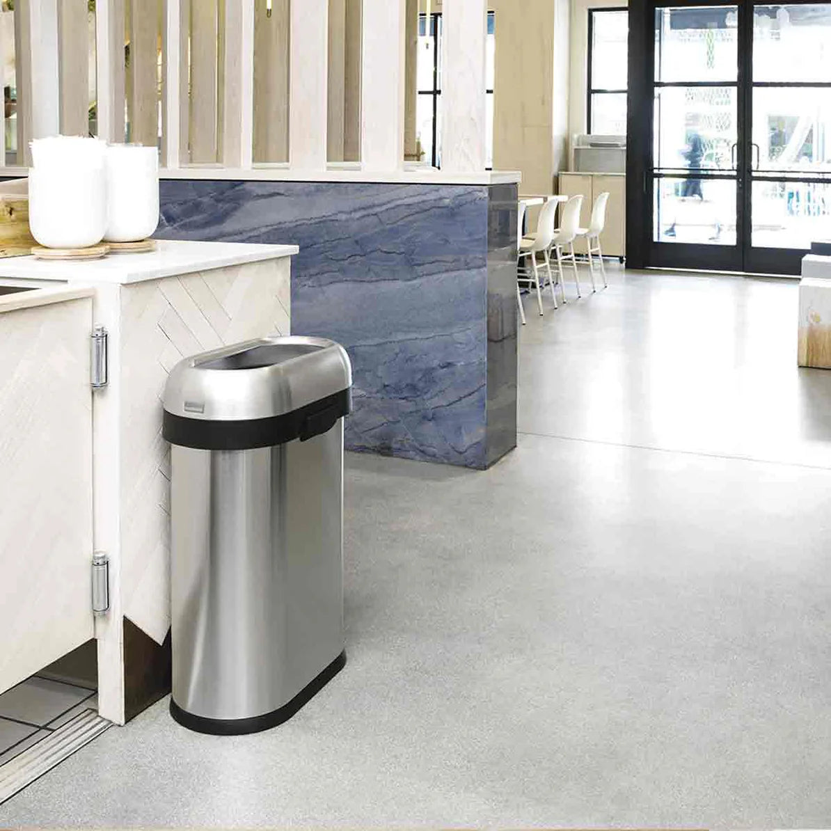 The image shows a sleek, stainless steel trash can positioned in a modern, open-concept kitchen or dining area. The trash can features a black, oval-shaped open-top lid, blending seamlessly with the contemporary decor. The setting includes light-colored wood accents and minimalist furnishings, creating a clean and spacious environment. The trash can's design complements the stylish interior, making it a functional yet attractive addition to the space.