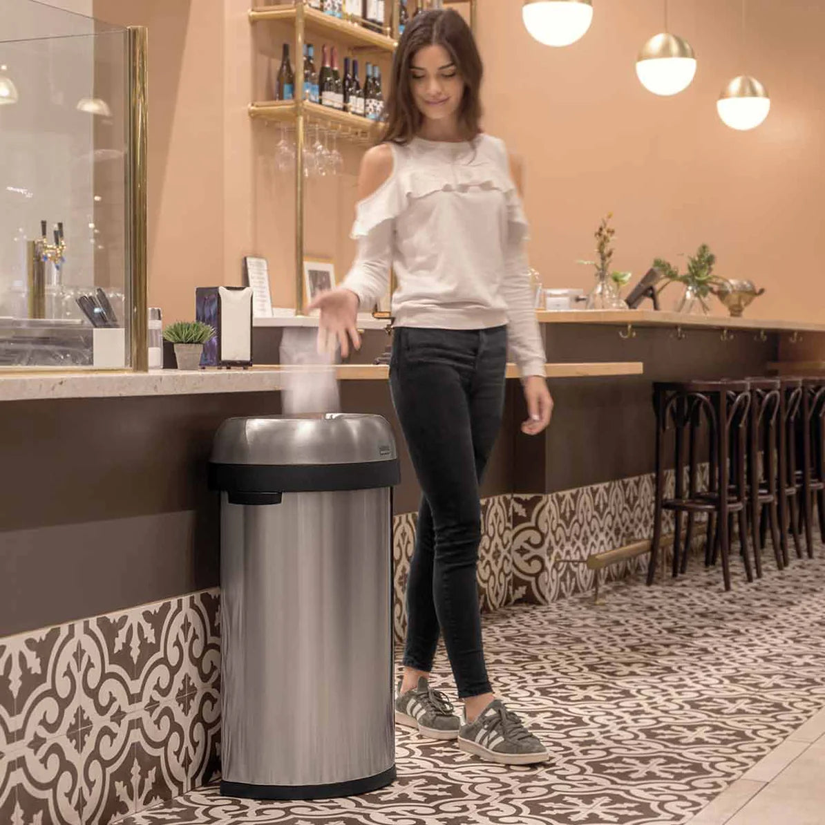 The image shows a woman using a sleek, stainless steel trash can in a stylish indoor setting, likely a cafe or restaurant. The trash can has a dome-shaped lid with a black trim and a foot pedal for hands-free operation. The setting features patterned flooring and modern decor, with the trash can blending seamlessly into the environment. The image highlights the practicality and elegance of the trash can, making it an attractive and functional choice for public spaces.