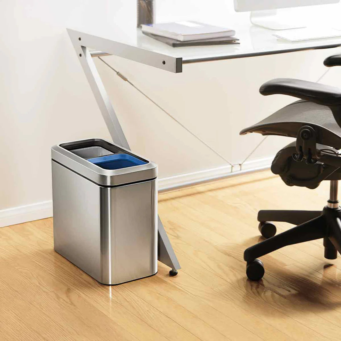 The image shows a modern office setup with a sleek, rectangular stainless steel trash can placed beside a glass-top desk. The trash can features a dual-compartment design, with one compartment having a blue liner visible at the top. The stainless steel finish and black accents give it a contemporary appearance, complementing the clean lines of the desk and the overall minimalist decor of the office. The compact size fits neatly beside the desk, making it convenient for waste disposal while maintaining a tid