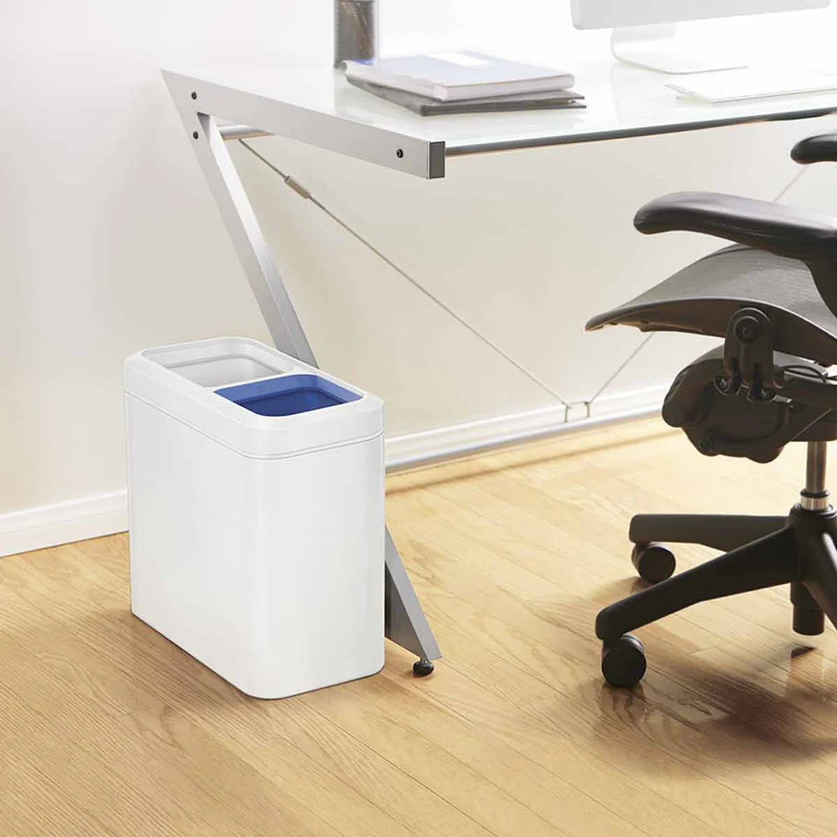 The image shows a modern office setting with a sleek, rectangular white trash can placed beside a glass-top desk. The trash can features a dual-compartment design, with one compartment having a blue liner visible at the top. The minimalist design of the trash can, with smooth, rounded edges and a clean white finish, complements the contemporary workspace. The compact size allows it to fit neatly beside the desk, making it convenient for waste disposal while maintaining a tidy and organized appearance. The o