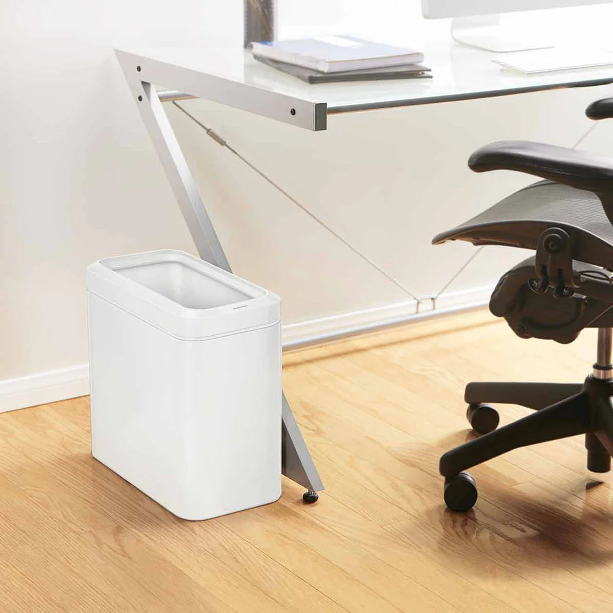 The image shows a modern office setup featuring a white rectangular trash can placed next to a sleek glass-top desk. The trash can has a minimalist design with a clean, smooth finish that complements the contemporary workspace. The compact size fits neatly beside the desk, making it convenient for disposing of waste without taking up much space. The office chair and light wood flooring add to the clean and organized atmosphere, with the white trash can seamlessly blending into the overall aesthetic. This se