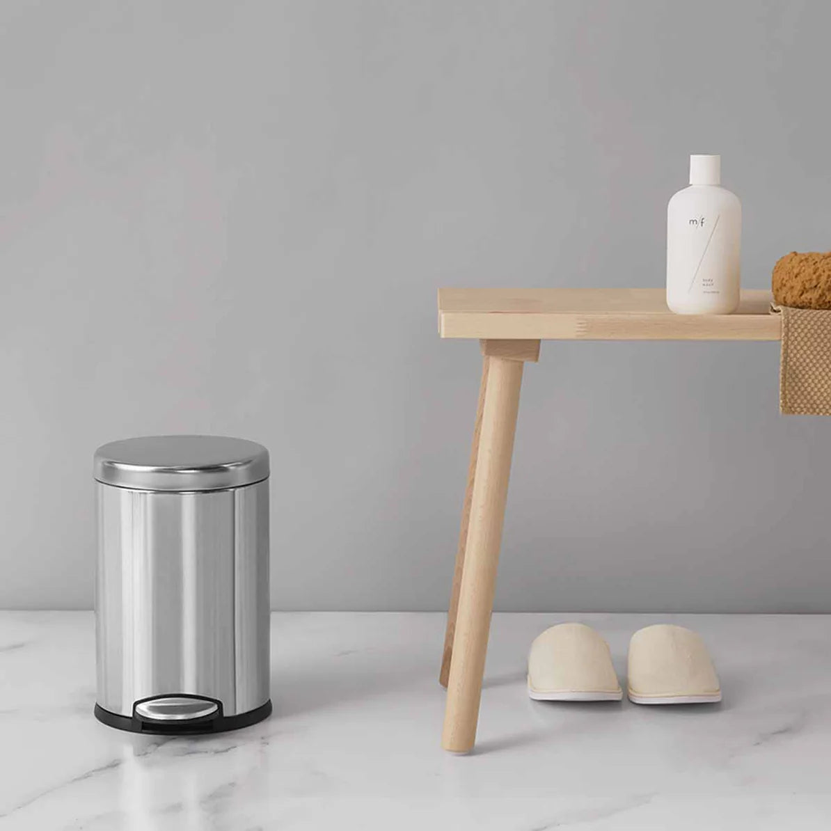 The image shows a small, stainless steel cylindrical trash can with a foot pedal for hands-free operation, placed on a clean, minimalist bathroom floor. The trash can features a polished finish with black accents at the base, adding a modern touch to the setting. Next to the trash can is a wooden bench with simple lines, holding a bottle of bath product and a loofah. A pair of white slippers are neatly placed under the bench, creating a serene and organized atmosphere. The overall scene conveys a sense of c