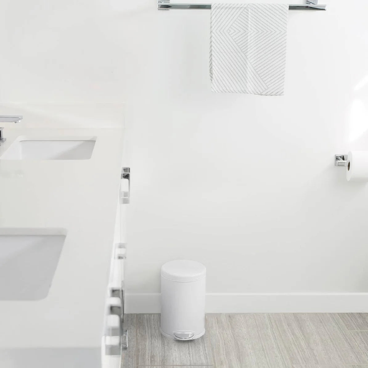 The image shows a minimalist bathroom with a small, white cylindrical trash can placed near the wall. The trash can features a foot pedal for hands-free operation and blends seamlessly with the clean, all-white decor of the bathroom. The surrounding area includes a white countertop with a sink and light-colored flooring, creating a bright and airy atmosphere. The compact size and sleek design of the trash can make it a subtle yet functional addition to the space, maintaining the room's modern and uncluttere