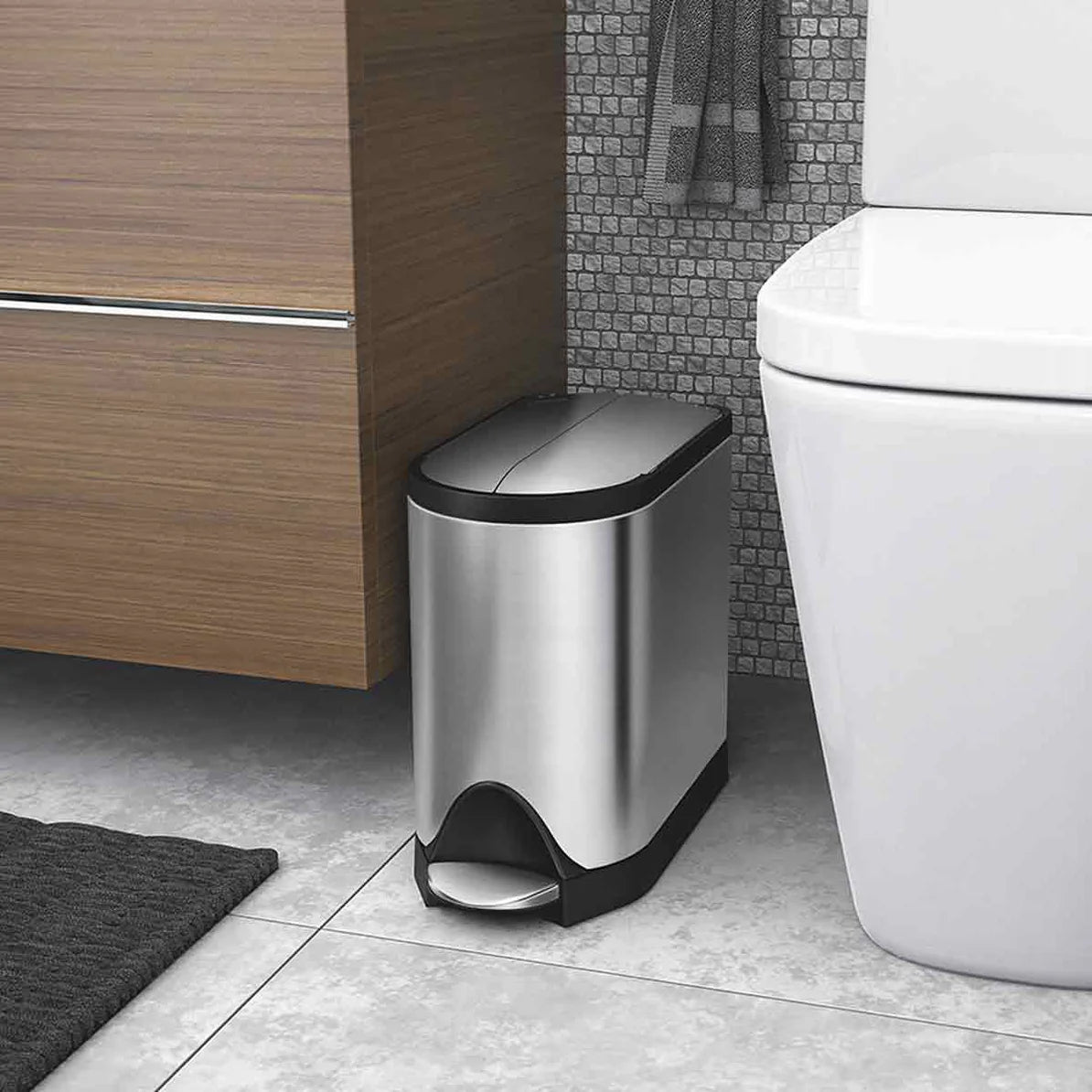 The image shows a sleek, stainless steel trash can with a modern design, positioned neatly in a bathroom beside a toilet. The trash can features a foot pedal for hands-free operation and a compact size that fits perfectly in small spaces. The design is minimalist, blending seamlessly with the contemporary decor of the bathroom.