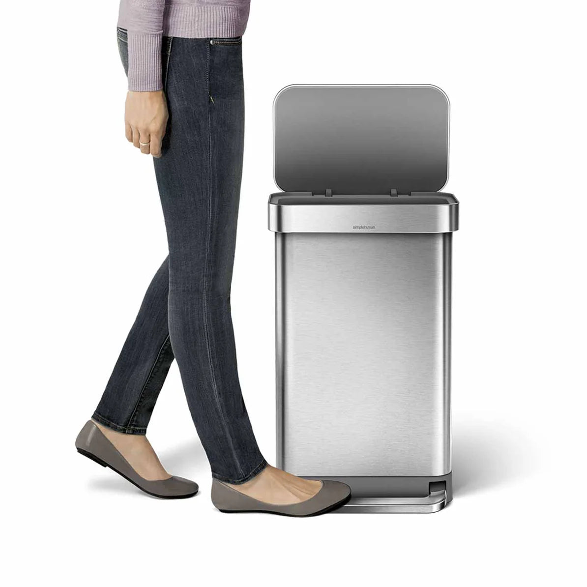 The image shows a person standing next to a Simplehuman trash can with a brushed stainless steel finish. The trash can has a rectangular shape with slightly rounded corners, and the lid is open, revealing the sleek interior. The person, dressed in jeans, flats, and a light purple sweater, is pressing the foot pedal at the base of the trash can, demonstrating its hands-free operation. The brushed stainless steel body of the trash can is complemented by a matching lid and foot pedal, giving it a modern and so