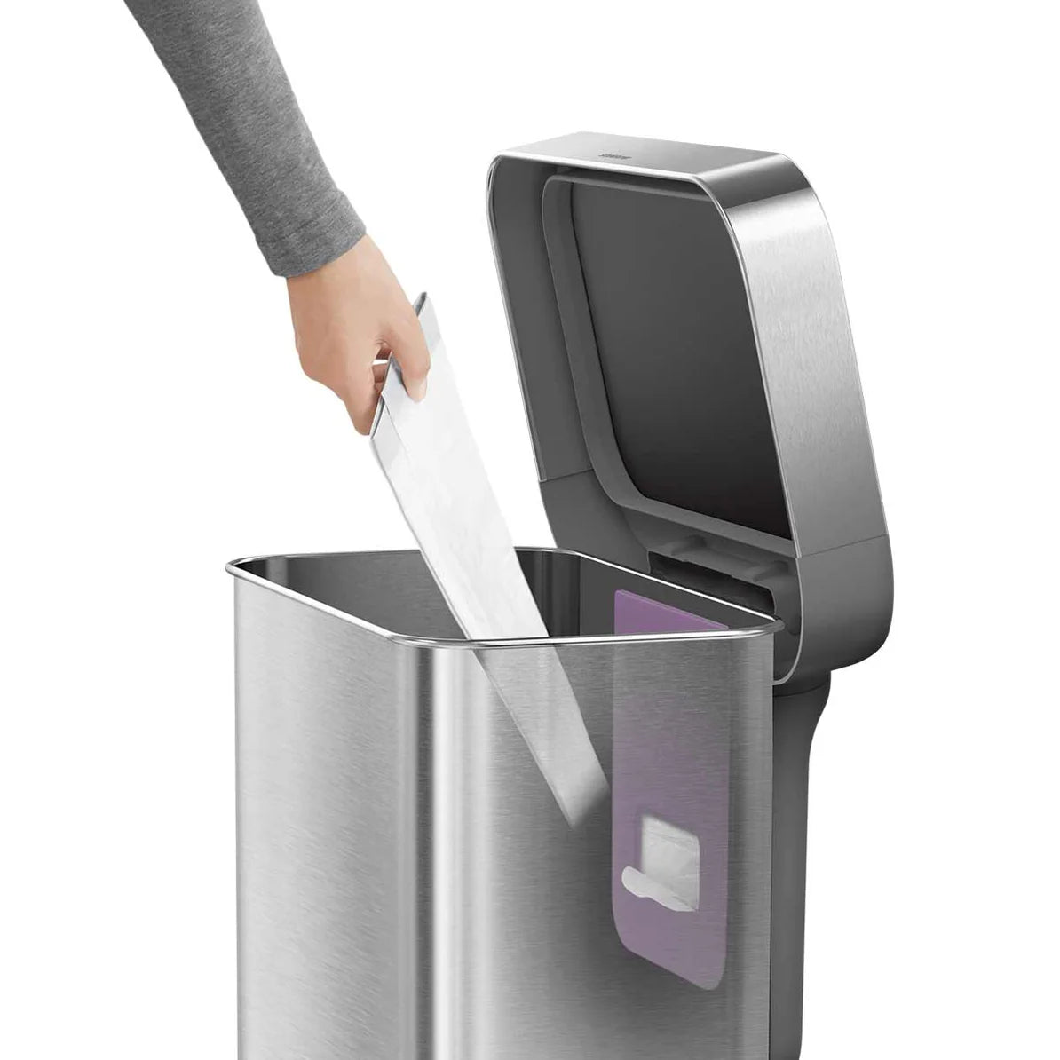 The image shows a person using a Simplehuman trash can with a brushed stainless steel finish. The lid is open, and the person, dressed in a gray sleeve, is placing an item, likely a piece of paper, into the trash can. Inside the can, a purple liner pocket is visible, which is designed to hold a roll of trash bags for easy access and quick changes. The brushed stainless steel exterior gives the trash can a sleek and modern look, while the interior liner pocket adds functionality by making it easier to manage