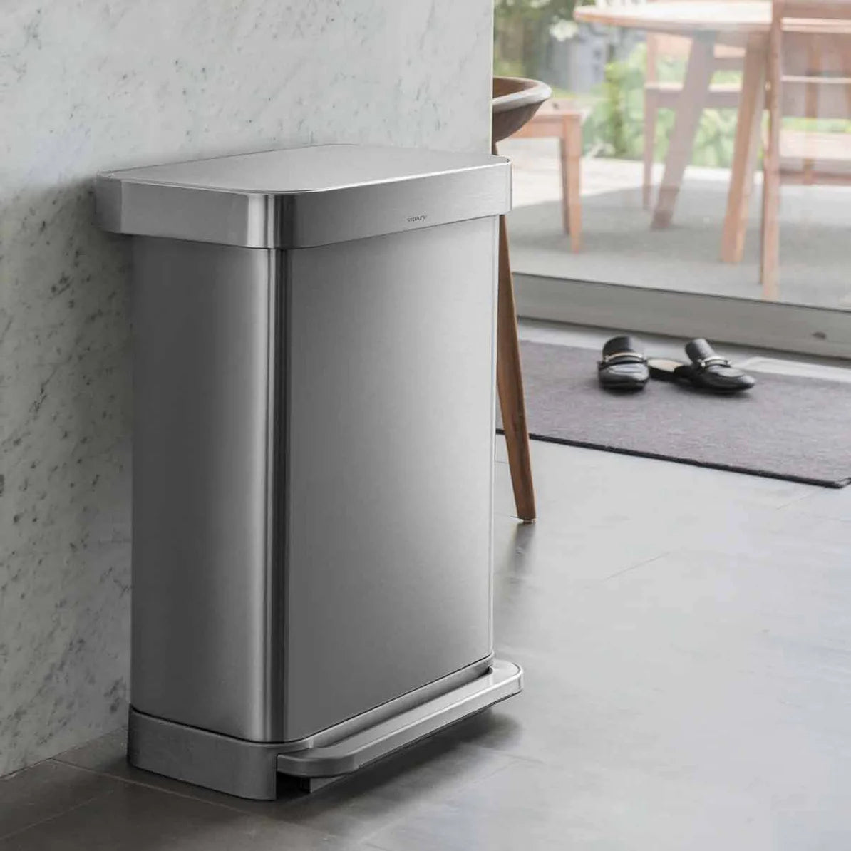 The image shows a Simplehuman trash can with a brushed stainless steel finish, positioned against a marble wall in a modern, minimalist setting. The trash can has a rectangular shape with slightly rounded corners, and the stainless steel exterior gives it a sleek, polished appearance. The foot pedal at the base, also in stainless steel, provides hands-free operation and matches the overall design. The can blends seamlessly with the high-end materials of the surroundings, including the marble wall and the mi