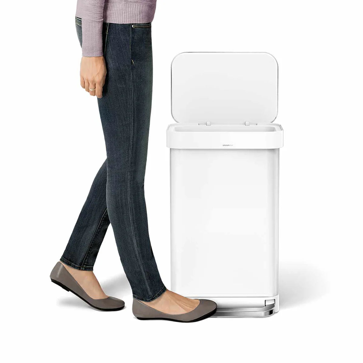 The image shows a person standing next to a white Simplehuman trash can. The trash can has a rectangular shape with a flat, open lid. The person, dressed in jeans, flats, and a light purple sweater, is using the foot pedal at the base of the trash can to open the lid, demonstrating the hands-free operation. The can has a sleek, modern design with a matte white finish, making it a stylish and functional addition to any home or office. The foot pedal is accented with a metallic finish, adding a touch of sophi