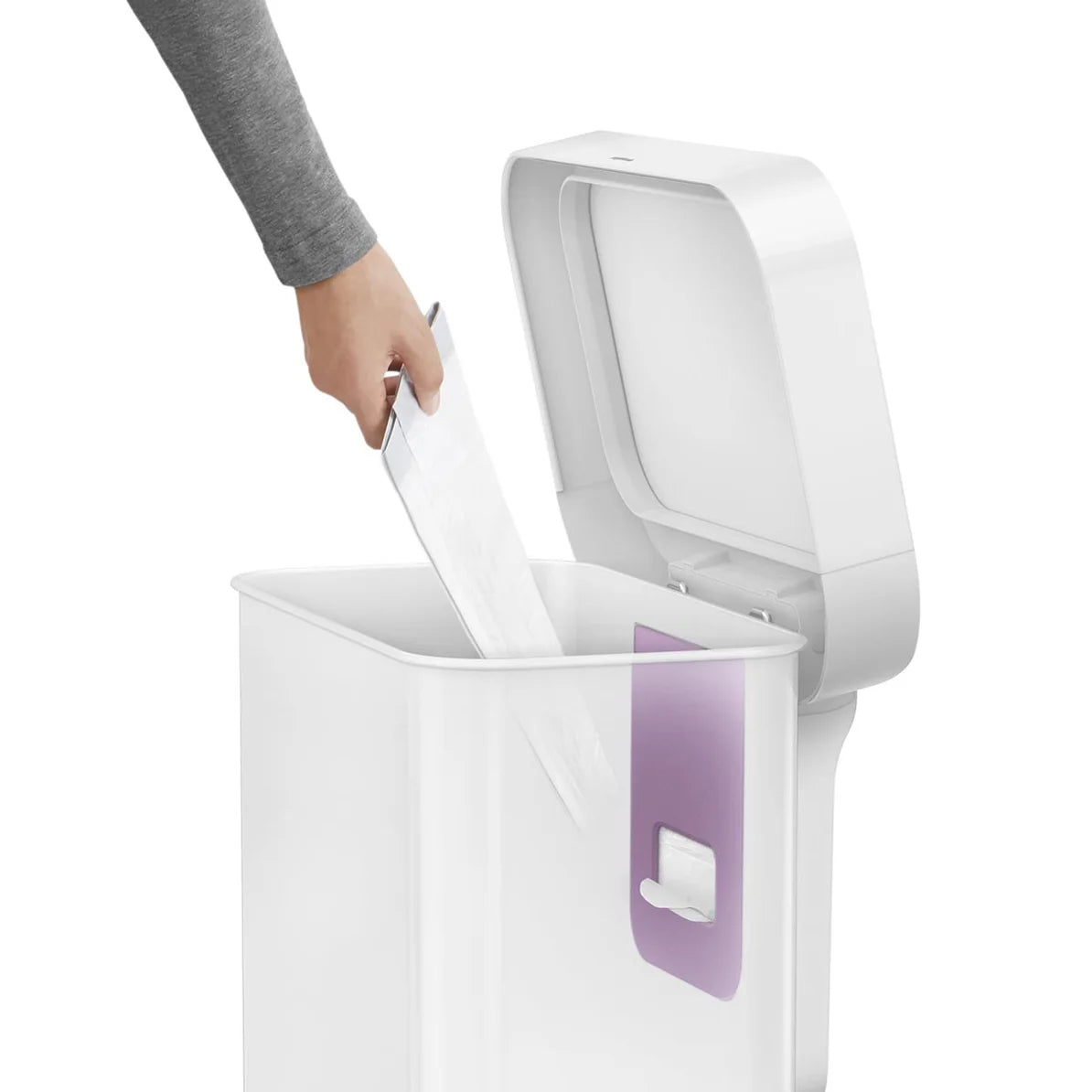 The image shows a person using a white Simplehuman trash can with an open lid. The person, dressed in a gray sleeve, is placing an item, likely a piece of paper, into the trash can. Inside the can, a purple liner pocket is visible, which holds a roll of trash bags, allowing for easy access and quick changes. The trash can has a sleek, modern design with a white finish that matches the lid and body. The lid is open, showing the clean and functional interior of the trash can, designed to make waste disposal m