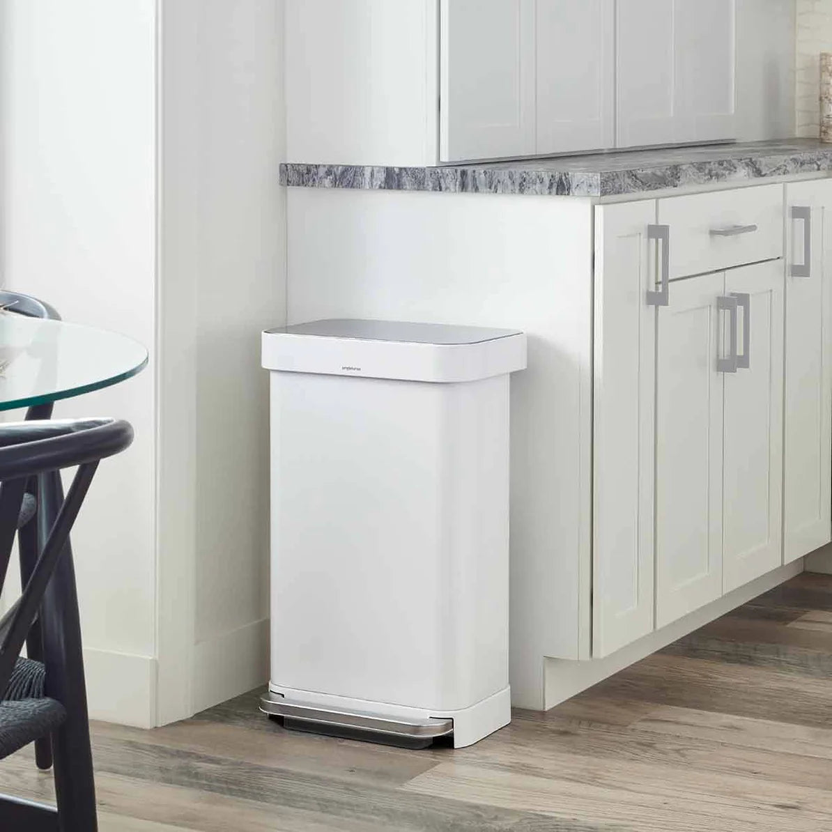 The image shows a white Simplehuman trash can placed in a modern kitchen setting. The trash can is positioned against a wall next to a cabinet with a marble countertop, seamlessly blending into the clean, contemporary design of the space. The can has a rectangular shape with slightly rounded corners and a stainless steel foot pedal at the base, allowing for hands-free operation. The white finish complements the white cabinetry, creating a cohesive and minimalist aesthetic. The overall design of the trash ca