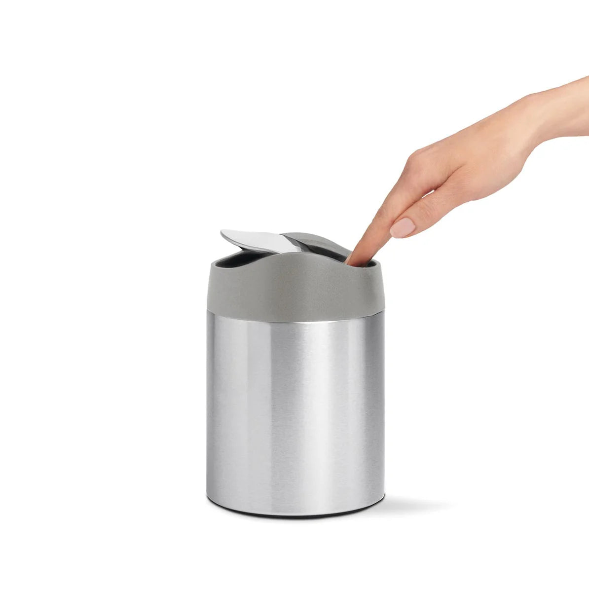 The image shows a Simplehuman mini round stainless steel trash can with a swing lid, being used by a person. The person is seen pressing down on the matte gray swing lid with their finger, demonstrating the easy and functional design of the trash can. The body of the trash can is made of brushed stainless steel, providing a sleek and modern look. This compact trash can is ideal for small spaces such as bathroom countertops, vanities, or desks. The swing lid is designed to open with a light touch and close a