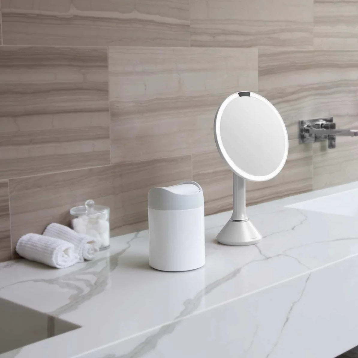 The image shows a stylish bathroom vanity setup featuring two Simplehuman products. On the left side of the countertop is a small Simplehuman trash can with a white body and a matte gray swing lid. The trash can is compact and has a modern, minimalist design, making it ideal for bathroom use. Next to the trash can is a Simplehuman sensor mirror, also with a sleek, modern design. The mirror has a circular shape and is mounted on a tall, conical stand, with integrated LED lighting that provides optimal illumi