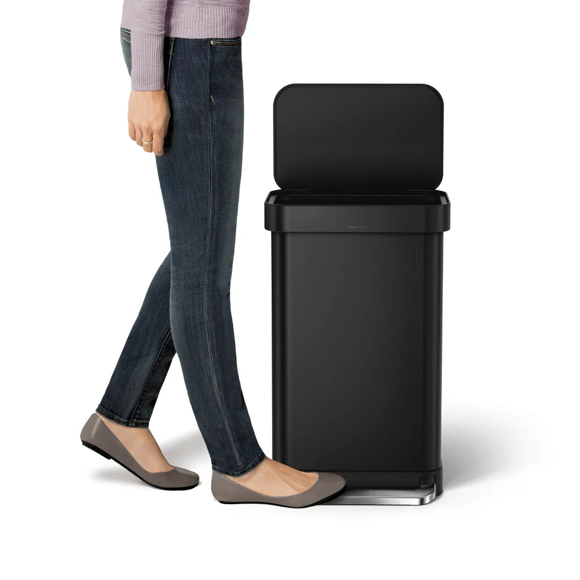 The image shows a person standing next to a black Simplehuman trash can. The trash can has a rectangular shape with a flat, open lid. The person, dressed in jeans, flats, and a light purple sweater, is using the foot pedal at the base of the trash can to open the lid, demonstrating the hands-free operation. The can has a sleek, modern design with a matte black finish, making it a stylish and functional addition to any home or office. The foot pedal is accented with a metallic finish, adding a touch of sophi