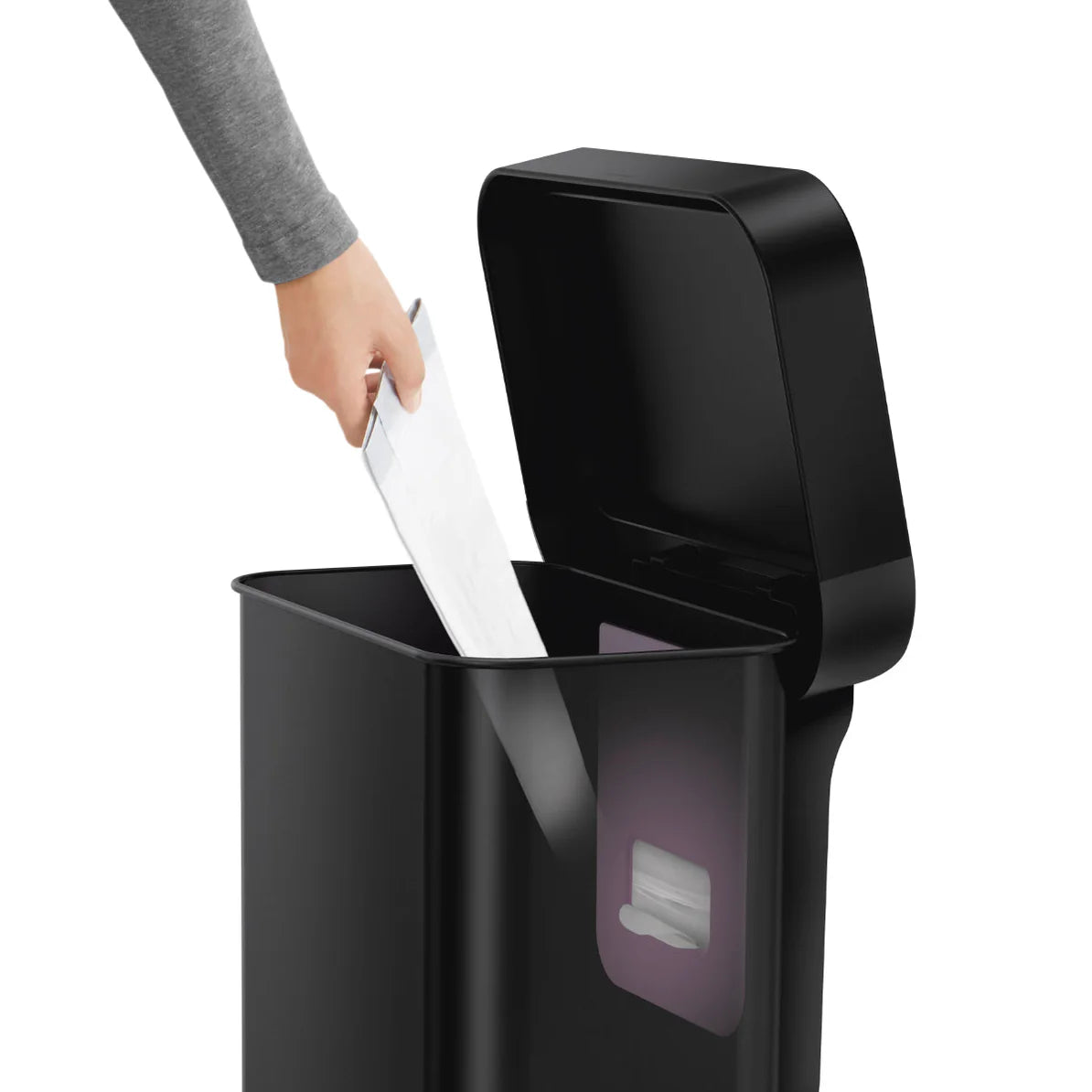 The image shows a person placing an item, likely a piece of paper, into a Simplehuman trash can with a black finish. The can has a sleek, modern design with a rectangular shape and rounded edges. The lid is open, revealing the spacious interior of the can. A liner pocket is visible inside the can, which is a feature in some Simplehuman models that allows for easy access to liner bags. The person's arm, dressed in a gray sleeve, is seen as they drop the paper into the trash can. This image highlights the can