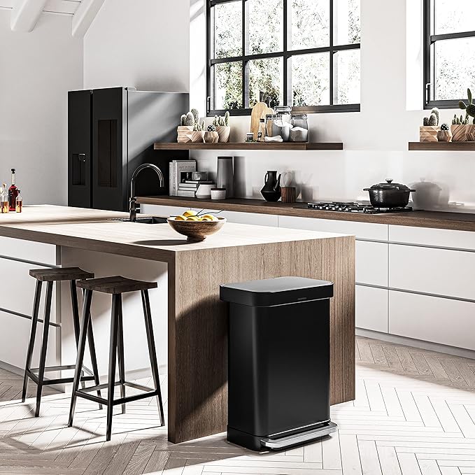 A modern kitchen setting featuring a black rectangular trash bin with a sleek design placed at the end of a wooden kitchen island. The kitchen has contemporary elements, such as white cabinetry, a large black refrigerator, and minimalist open shelves adorned with kitchen essentials and potted plants. The large windows allow natural light to flood the space, highlighting the harmonious blend of wood and modern black accents.