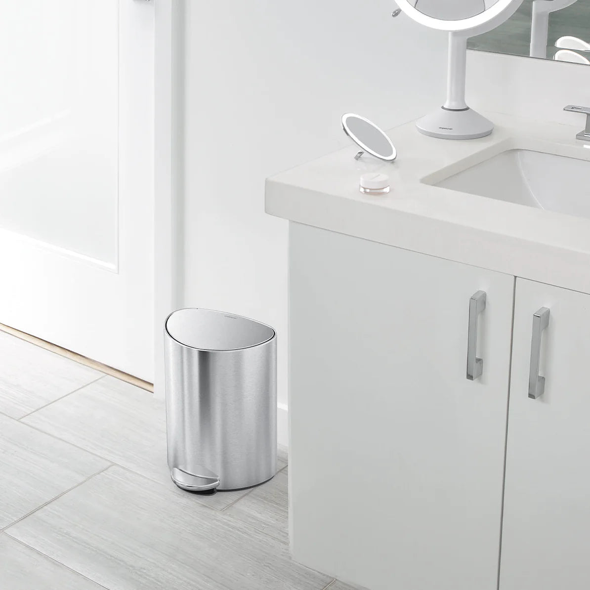 The image depicts a modern bathroom setting with a stainless steel trash can placed near the corner, next to a white bathroom vanity with a sink. The trash can has an oval shape with a foot pedal at the front for hands-free operation. The overall bathroom decor is clean and minimalist, featuring light-colored tiles and a white countertop. On the countertop, there is a mirror with a stand and a few small items, reinforcing the sleek and organized aesthetic of the space. The trash can's design complements the