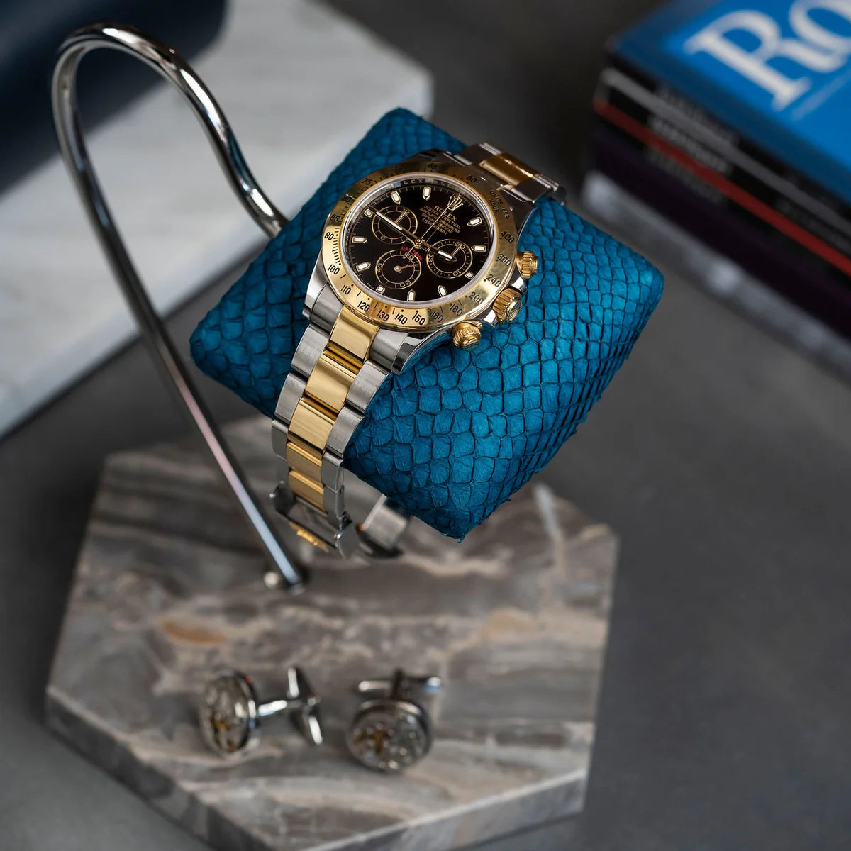 A luxurious wristwatch with a two-tone metal band (gold and silver) and a black dial with gold accents is displayed on a blue textured cushion. The cushion is mounted on a sleek, curved metal stand with a hexagonal gray and brown veined marble base. The setup is placed on a dark surface, with a pair of cufflinks positioned nearby. In the background, there are books, adding a sophisticated and elegant touch to the overall presentation.