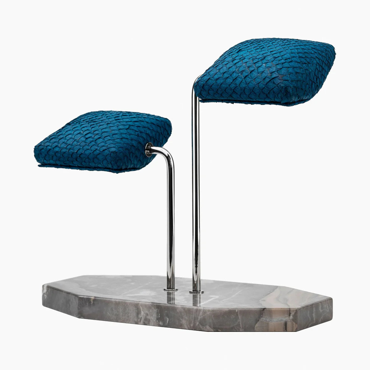  A display stand featuring two blue textured cushions mounted on elegant silver curved supports. The base of the stand is a gray marble slab with subtle veining and an irregular shape. The design is sophisticated and luxurious, ideal for showcasing wristwatches or similar items. The vibrant blue cushions contrast beautifully with the polished silver supports and marble base, creating a refined and stylish presentation. The stand is shown from a slightly angled side view.