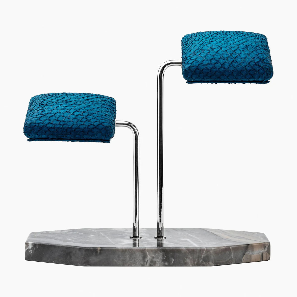 A display stand featuring two blue textured cushions mounted on elegant silver curved supports. The base of the stand is a gray marble slab with subtle veining and an irregular shape. The design is sophisticated and luxurious, ideal for showcasing wristwatches or similar items. The vibrant blue cushions contrast beautifully with the polished silver supports and marble base, creating a refined and stylish presentation.