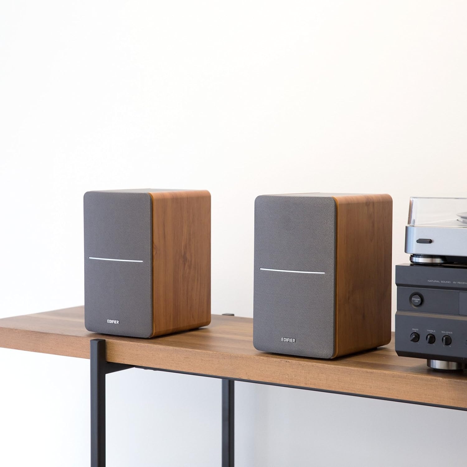 A pair of wooden bookshelf speakers with grey fabric grilles, elegantly placed on a wooden shelf alongside an audio system, offering a minimalist and sophisticated home audio setup.