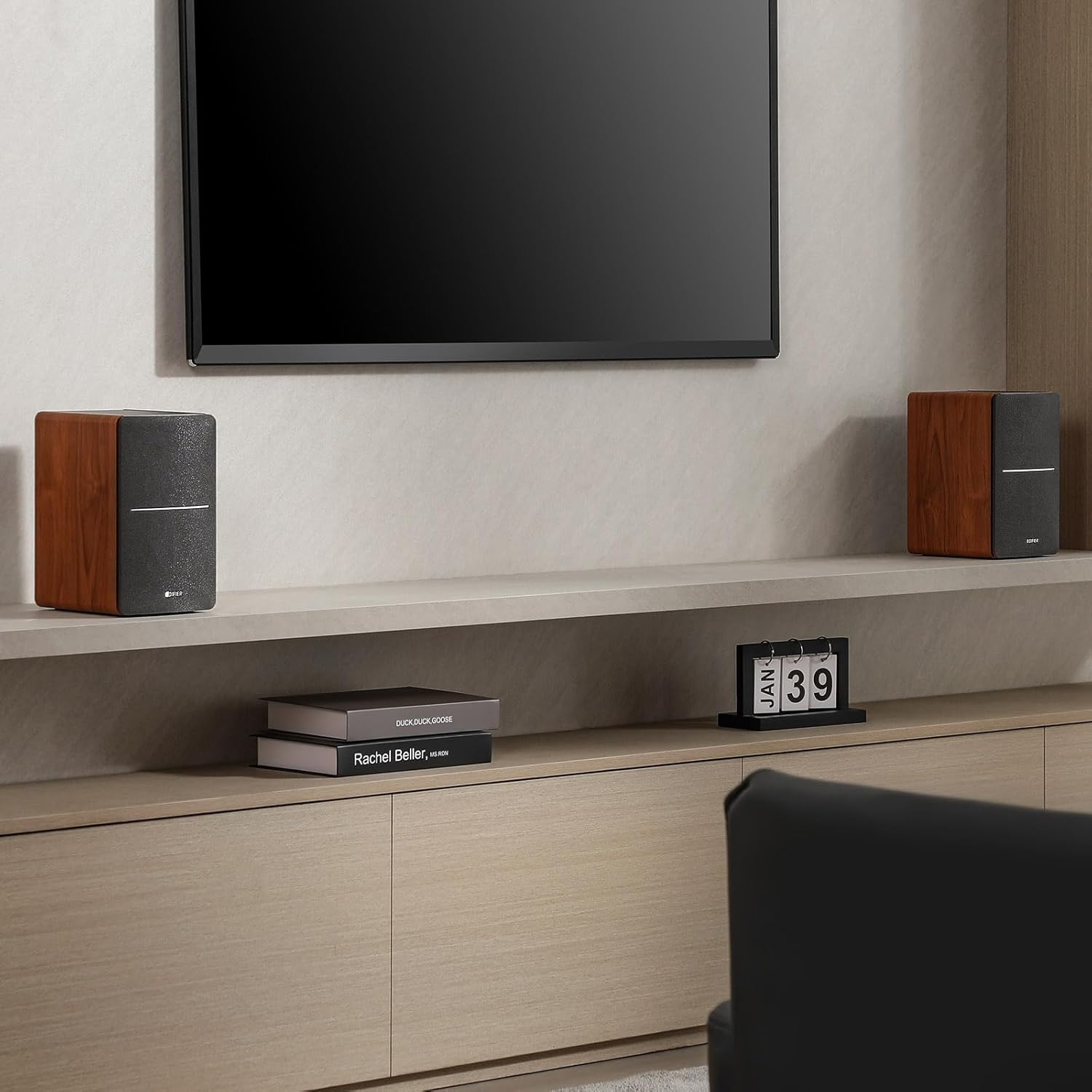 This image showcases a modern living room setup with a sleek wall-mounted television as the focal point. Below it, a minimalist shelf holds a pair of compact bookshelf speakers with a wooden and gray finish, flanking the TV. The cabinet below adds to the tidy aesthetic, featuring neatly arranged books and a small calendar. This design combines style and functionality, creating a sophisticated entertainment space. Let me know if you need further assistance!