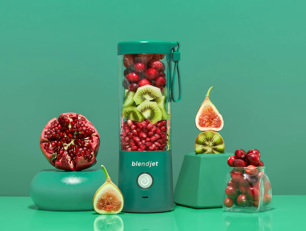 A vibrant green BlendJet blender packed with kiwis, cranberries, and pomegranate seeds, surrounded by fresh fruits like a split pomegranate, figs, and cherries in a glass jar, set against a matching green background.