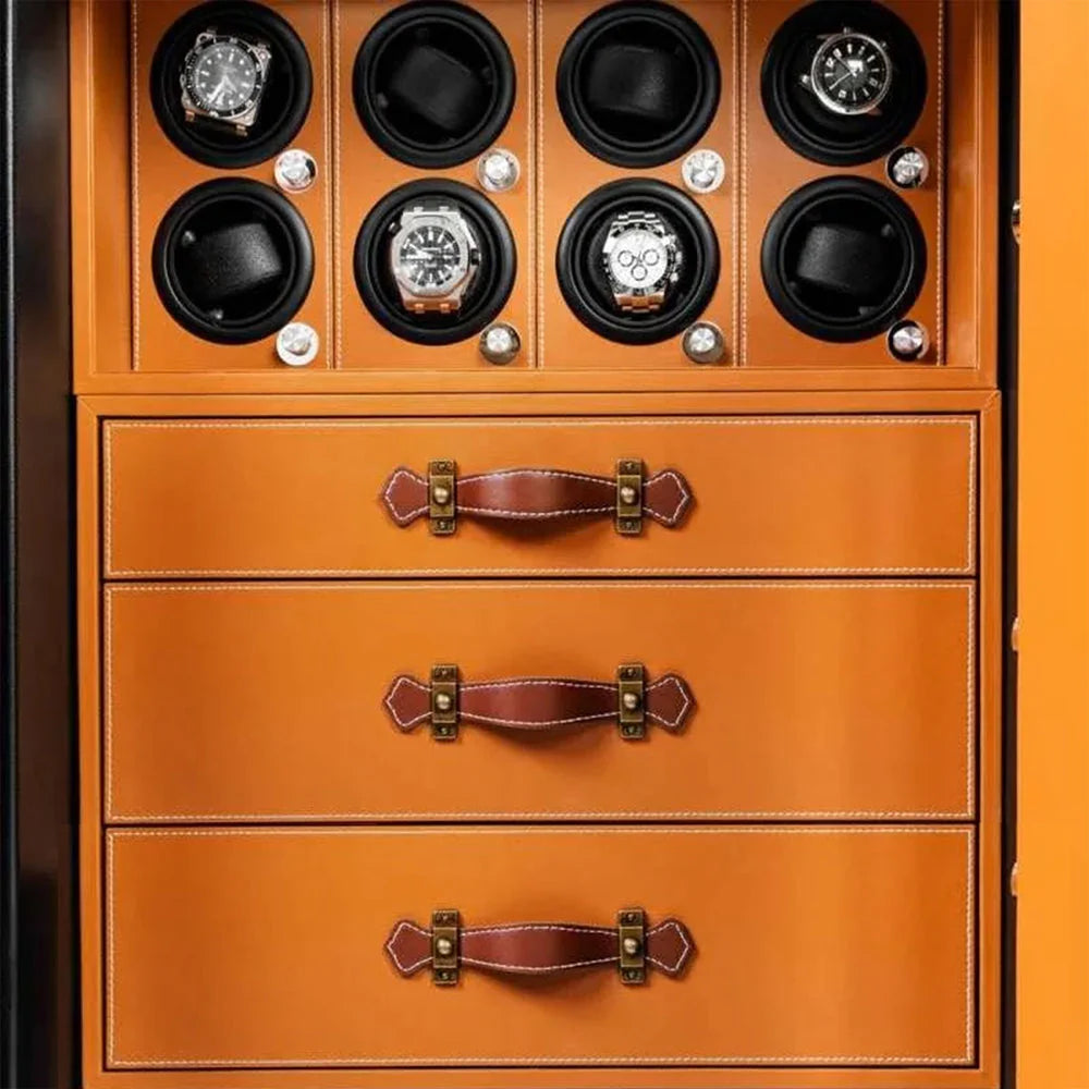  The interior of a safe featuring three orange drawers with brown leather handles and white stitching. Above the drawers, there are eight circular watch winders, each containing a luxury watch, designed to keep the watches running when not being worn. The setup displays a combination of elegant craftsmanship and practical storage.