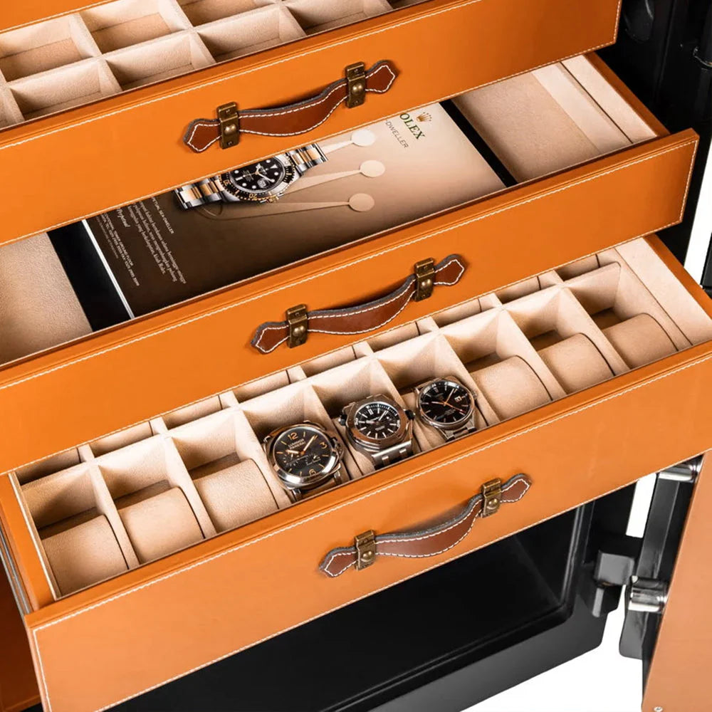 Close-up view of three orange drawers inside a safe, each with leather handles and white stitching. The top two drawers have multiple compartments for storing small items, while the bottom drawer contains several watches, neatly arranged in individual slots. An open brochure featuring a luxury watch is partially visible in the top drawer.