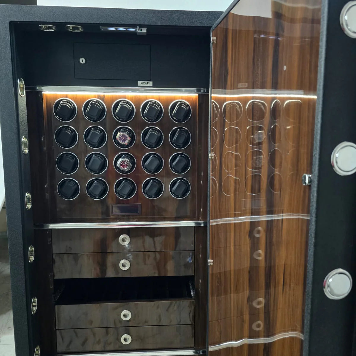  Interior view of a luxurious safe with a high-gloss wood-grain finish. The top section is equipped with eighteen watch winders arranged in three rows, some holding watches. Below the watch winders are two closed drawers and one open drawer with metallic handles for additional storage. The interior of the door matches the wood-grain finish and has visible locking bolts, showcasing a blend of elegance and security. The safe is well-lit, highlighting its sophisticated design.