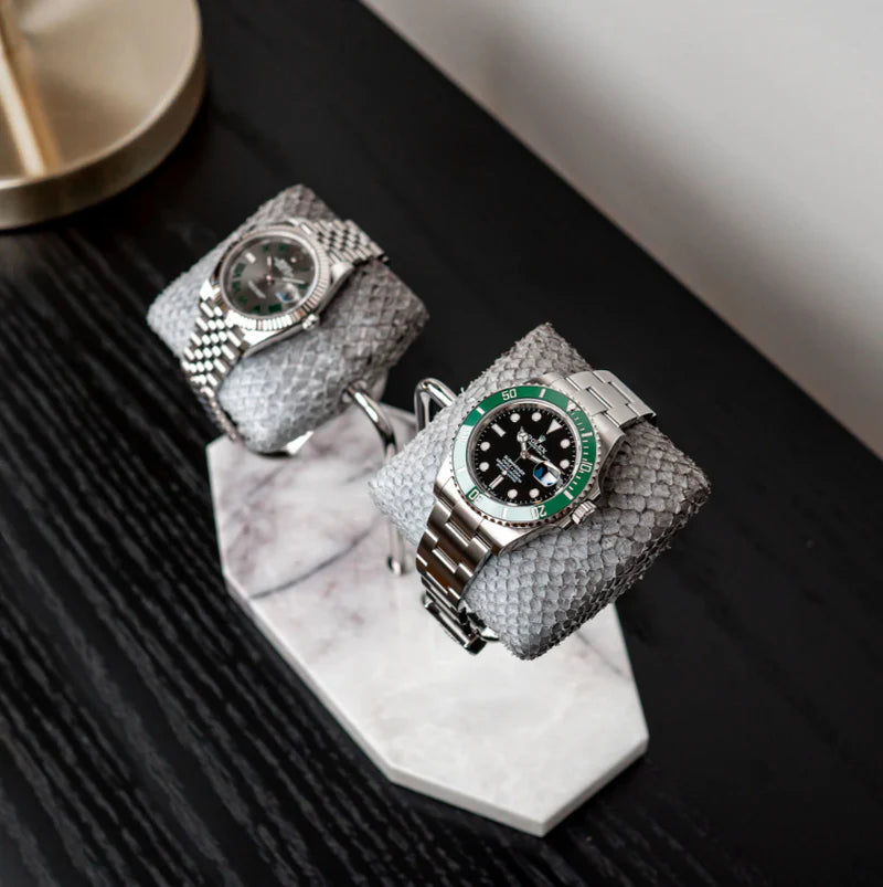 Two luxurious wristwatches are displayed on gray textured cushions, mounted on a double-arm stand with a white marble base. One watch has a silver metal band with a silver dial, while the other has a silver metal band with a black dial and green bezel. The display is set on a dark wooden surface, with a gold lamp partially visible in the background, creating a sophisticated and elegant presentation.