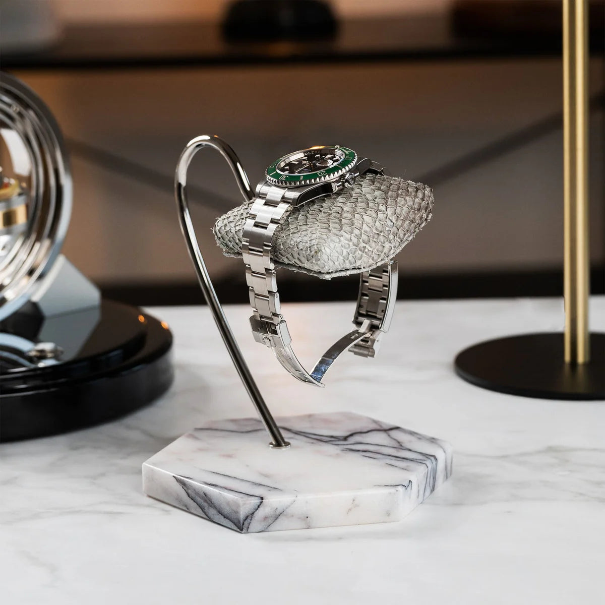  A wristwatch with a silver metal band and a green bezel is displayed on a textured gray cushion. The cushion is attached to a sleek, curved metal stand with a white marble base featuring gray veining. The display is set on a white marble surface, and the background includes modern decor elements, such as another watch display and a gold lamp, adding to the elegant and sophisticated ambiance.