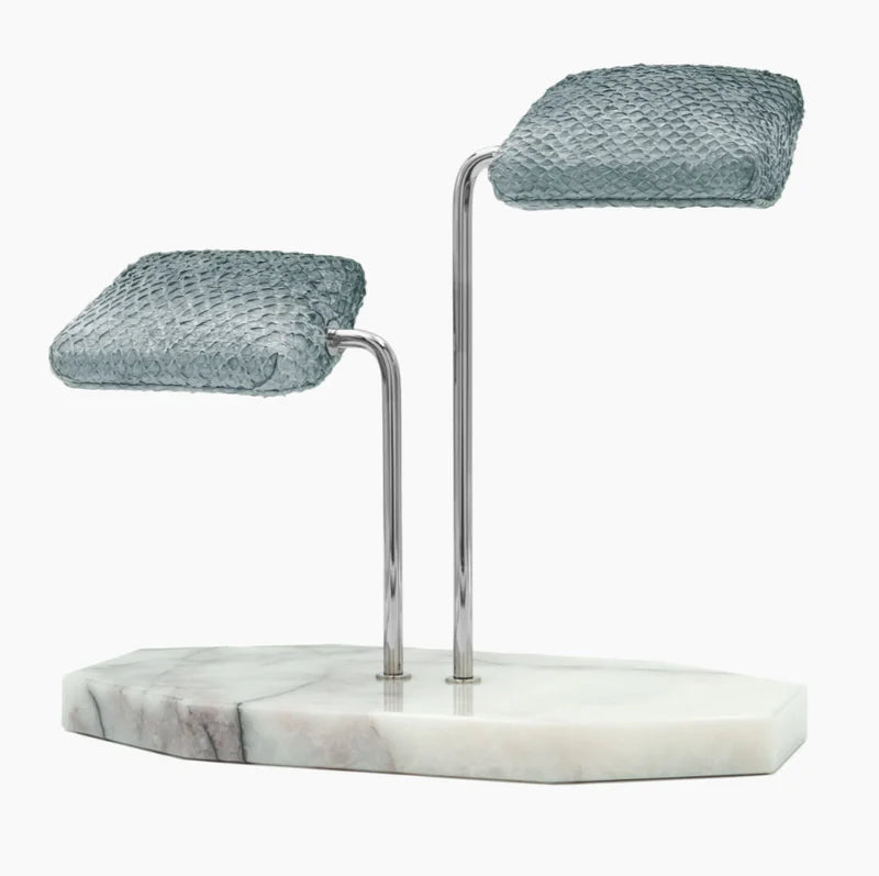  A display stand featuring two gray textured cushions mounted on elegant silver curved supports. The base of the stand is a white marble slab with subtle gray veining and an irregular shape. The design is sophisticated and luxurious, ideal for showcasing wristwatches or similar items. The combination of the gray cushions, silver supports, and marble base creates a refined and stylish presentation.