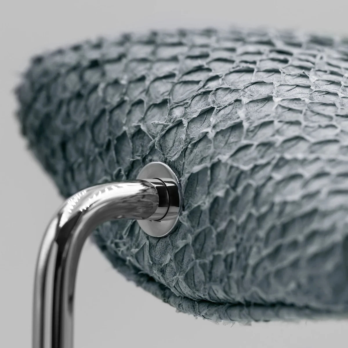 A close-up view of a gray textured cushion attached to a shiny silver curved metal support. The cushion has a scaly, reptile-like texture, and the metal support is secured with a polished silver screw. The image highlights the luxurious materials and the detailed craftsmanship of the display stand.