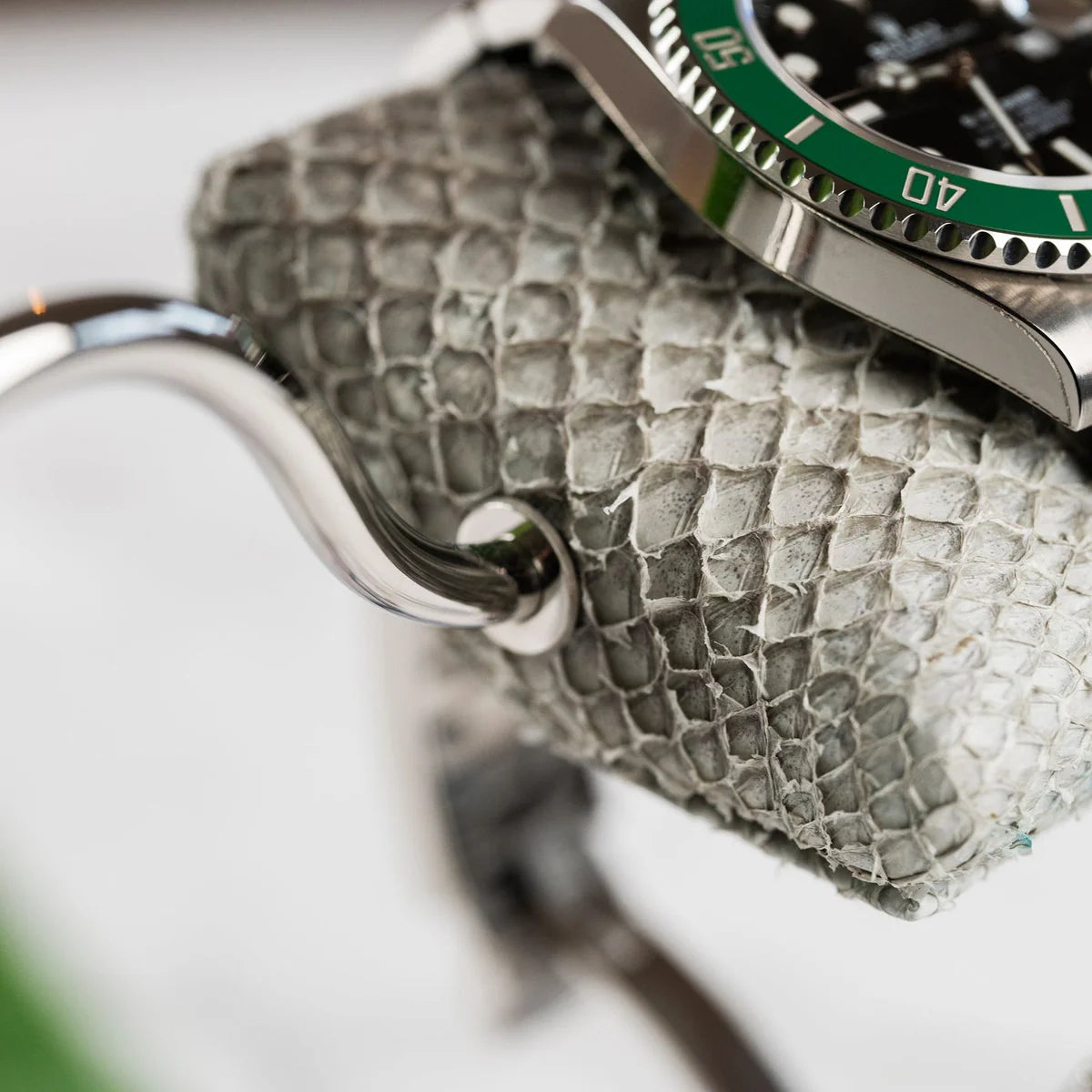 A close-up view of a wristwatch with a green bezel and silver metal band displayed on a textured gray cushion. The cushion is attached to a shiny curved metal stand, with the attachment point clearly visible. The texture of the cushion and the polished surface of the watch and stand are highlighted, emphasizing the luxurious and detailed design of the display setup.