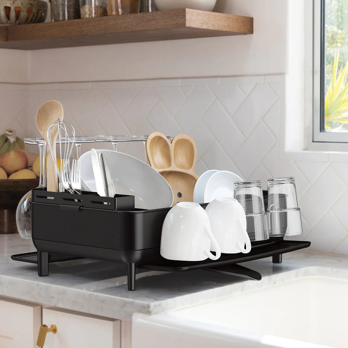 The image shows a modern kitchen scene with a black dish drying rack placed on a marble countertop next to a sink. The dish rack is well-organized, holding several clean dishes, including plates, bowls, cups, and glasses. There is also a section in the rack dedicated to utensils like spoons and spatulas. The drying rack has a sleek design with raised legs, allowing for proper drainage. The background features a white tiled backsplash, wooden shelving with various kitchen items, and a window that brings in n