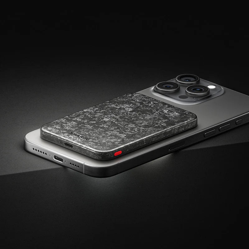 A sleek, textured gray MagSafe-compatible accessory, resembling a power bank or wallet, attached to the back of a modern smartphone with triple rear cameras. The accessory has a stone-like finish with a small red indicator light on the side, complementing the phone's metallic design. The setup is placed on a dark background, emphasizing the premium and minimalist aesthetic.