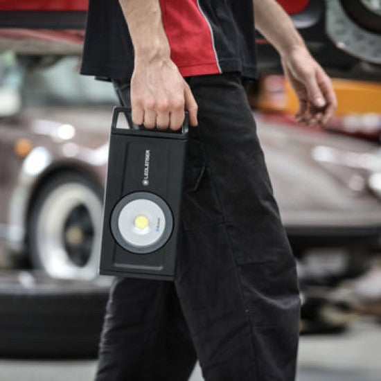 A person carrying a LEDLenser portable light by its handle. The light is black with a circular LED in the center, and the person is standing in what appears to be a garage or workshop, with a car visible in the background.