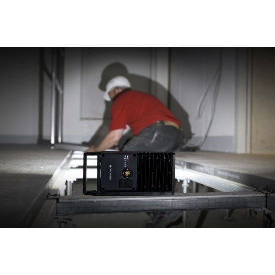  A LEDLenser portable light positioned on a metal surface, illuminating a workspace where a person in a red shirt and white hard hat is working. The worker, seen from behind, is focused on a task in a dimly lit environment, with the light providing essential visibility.