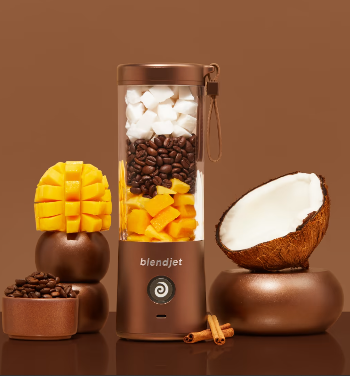 This is a BlendJet portable blender in a bronze hue, filled with layers of diced coconut, coffee beans, and mango chunks. On the left, there's a carefully carved mango displayed over bronze bowls containing coffee beans. On the right, a halved coconut rests on a bronze stand with cinnamon sticks accenting the scene. The entire composition exudes a warm, tropical feel with a luxurious twist, thanks to the cohesive brown tones and bronze highlights.