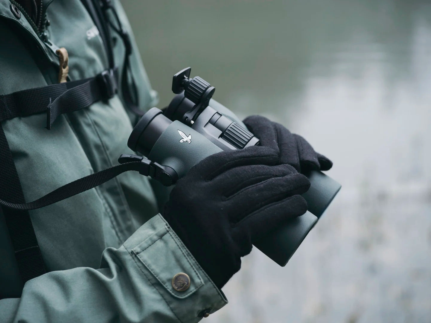 A person wearing a green jacket and black gloves holds a pair of Swarovski binoculars, emphasizing their ergonomic design and outdoor usability.