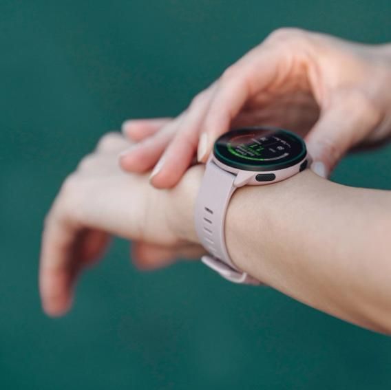  A close-up of a person wearing a Polar Pacer smartwatch on their left wrist. The person is adjusting or interacting with the watch using their right hand. The smartwatch screen is visible, displaying fitness tracking information. The watch has a purple strap with a buckle closure. The background is blurred, highlighting the watch and the person's hands.