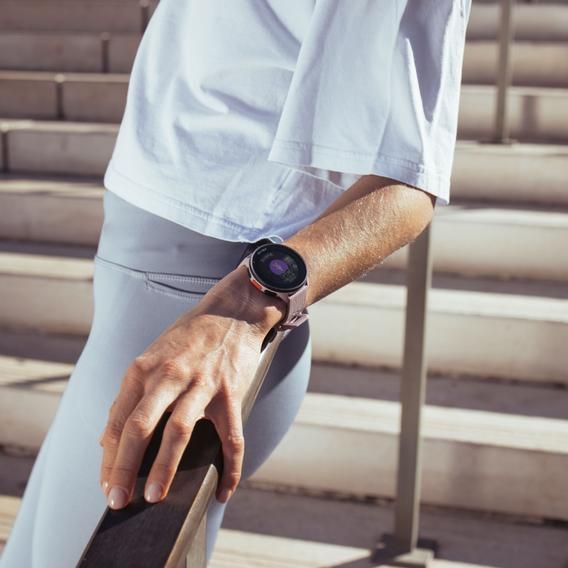 A person wearing a Polar Pacer smartwatch on their left wrist. The person is dressed in light-colored activewear, leaning on a handrail with stairs in the background. The smartwatch screen is visible, showcasing its fitness tracking capabilities. The watch has a purple strap, matching the person's sporty and casual outfit.