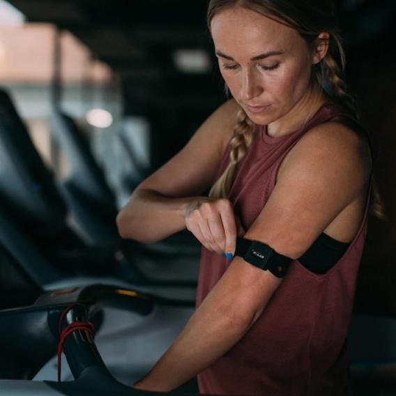 A woman adjusting a Polar optical heart rate sensor on her upper arm, secured with a black adjustable strap. She is in a gym setting, dressed in a sleeveless maroon top and braids, and is standing next to a piece of exercise equipment