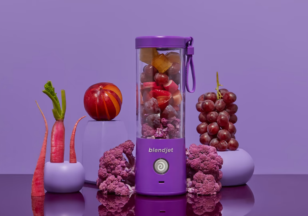 A portable purple BlendJet blender filled with frozen fruits and vegetables, surrounded by fresh produce like red carrots, a sliced apple, grapes, and purple cauliflower, set on a reflective surface against a monochromatic purple background.