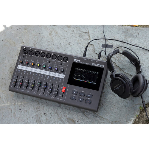 Zoom R20 audio mixer and recorder placed on a stone surface, accompanied by a pair of black headphones connected to the device. The mixer features multiple channels with faders, knobs, and a digital display screen showing audio levels. This setup highlights the portability and outdoor usability of the audio equipment, ideal for on-the-go recording and mixing.