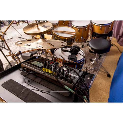 A drum set setup in a music studio, featuring multiple drums, cymbals, and a stool. In the foreground, there is a Zoom audio mixer and recorder on a stand, connected with various cables and wires. The scene captures a professional recording environment, emphasizing the integration of audio equipment for capturing drum performances. The setup highlights the detailed arrangement of drums and the use of advanced recording technology.