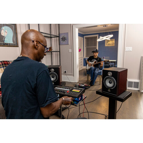 Two musicians in a home studio setting. The musician in the foreground is operating a Zoom audio mixer and recorder, adjusting controls with focused intent. The musician in the background is seated, playing an electric guitar, with studio monitors positioned around them. The room features modern decor, with musical equipment and cables set up for recording or practice. The scene captures a collaborative and creative atmosphere, highlighting the use of professional audio equipment in a home environment.