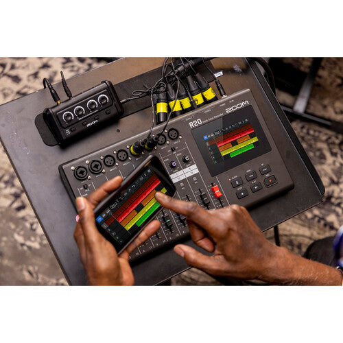 An overhead view of a person using a Zoom R20 audio mixer and recorder. The person's hands are visible, one hand adjusting controls on the mixer and the other holding a smartphone displaying the mixer's interface. The mixer is connected to various cables and an external device, likely a wireless receiver, indicating an active recording or mixing session. The scene is set on a tabletop with a patterned background.