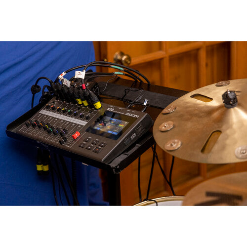 A close-up shot of an audio mixer with multiple cables connected, positioned next to a cymbal. The mixer displays various settings and levels, indicating it is actively being used to control and record audio. The background features wood paneling and part of a drum set.