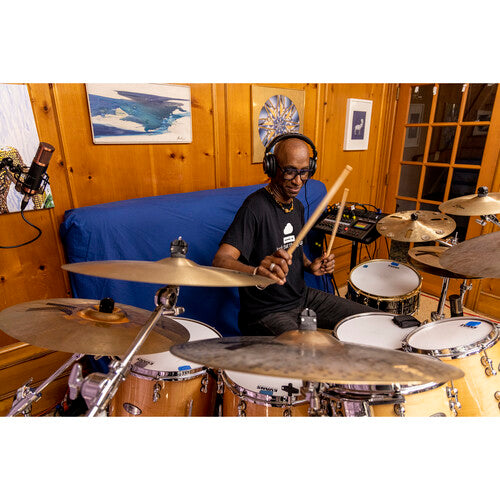 - A drummer wearing headphones is playing a drum set in a cozy, wood-paneled room. The room is adorned with framed pictures on the walls, and a microphone is set up near the drum set, capturing the performance.